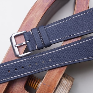 Puede incluir: Correa de reloj de cuero azul marino con costuras blancas. La correa tiene una hebilla plateada y se muestra sobre una superficie de madera. El cuero tiene un aspecto texturizado.