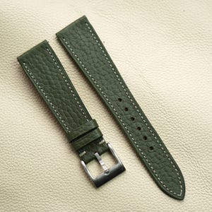 Peut inclure: Bracelet de montre en cuir vert olive avec surpiqûres blanches. Le bracelet a une surface texturée et une boucle argentée. Le bracelet est présenté sur une surface en cuir beige clair.