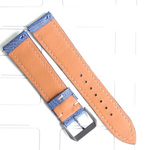 May include: Two-piece watch strap with a light blue textured top layer and a tan leather underside. The strap features a silver-colored buckle and is stitched with a light tan thread. The watch band is shown on a white surface.
