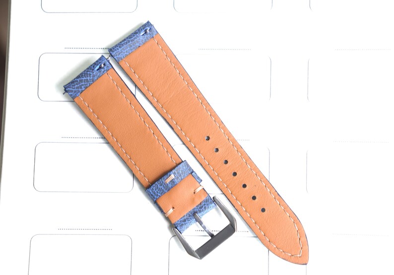 May include: Two-piece watch strap with a light blue textured top layer and a tan leather underside. The strap features a silver-colored buckle and is stitched with a light tan thread. The watch band is shown on a white surface.