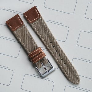 Puede incluir: Correa de reloj de dos piezas, de lona beige y cuero marrón. La correa tiene una hebilla plateada y presillas de cuero marrón, cosidas con hilo blanco. La correa del reloj está sobre una superficie blanca.