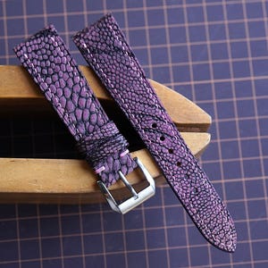 Puede incluir: Correa de reloj de dos piezas con un patrón púrpura texturizado y costuras blancas. La correa tiene una hebilla plateada y se muestra sobre una superficie de madera. Un elegante accesorio de reloj.