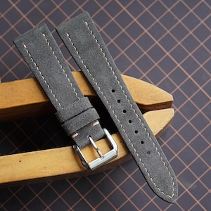 May include: Two gray suede watch straps with cream stitching and a silver buckle. The straps are displayed on a wooden clamp against a dark background with a grid pattern. The straps are new.