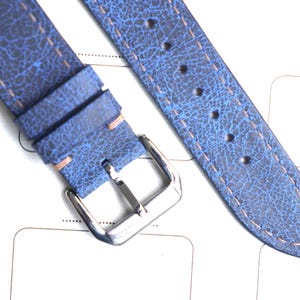 May include: Close-up of a blue leather watch strap with a textured finish. The strap has a silver buckle and light-colored stitching. The leather has a distressed appearance. The strap is lying on a white surface.