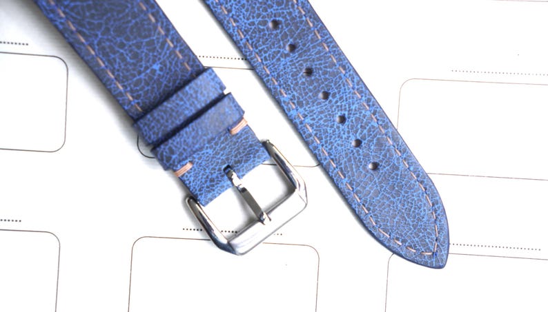 May include: Close-up of a blue leather watch strap with a textured finish. The strap has a silver buckle and light-colored stitching. The leather has a distressed appearance. The strap is lying on a white surface.
