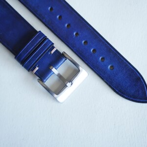 May include: Close-up of a blue leather watch strap with a silver buckle. The strap has several holes for adjustment and is made of a textured leather material. The watch band is a deep shade of blue.