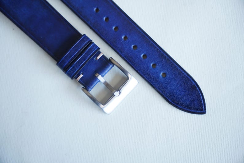 May include: Close-up of a blue leather watch strap with a silver buckle. The strap has several holes for adjustment and is made of a textured leather material. The watch band is a deep shade of blue.