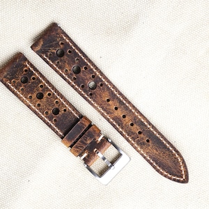 Op de afbeelding: Twee bruin lederen horlogebandjes met witte stiksels en meerdere ronde perforaties. Eén bandje heeft een zilverkleurige gesp. De bandjes zijn voor een horloge en liggen op een neutrale achtergrond.