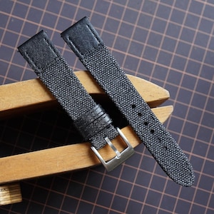 May include: A two-piece watch strap featuring a dark gray textured canvas material with black leather accents at the attachment points. The strap includes a silver-toned metal buckle and two matching black leather keepers for securing the band.