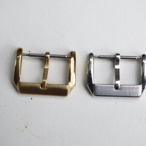 May include: Two watch buckles are displayed side-by-side. One is gold-toned, the other is silver-toned. Both have a rectangular frame with a central bar for the watch strap. The buckles are set against a white background.