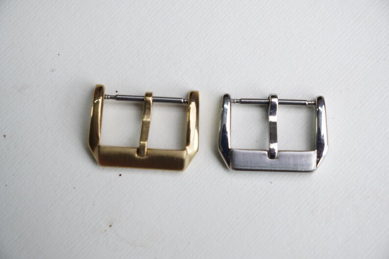 May include: Two watch buckles are displayed side-by-side. One is gold-toned, the other is silver-toned. Both have a rectangular frame with a central bar for the watch strap. The buckles are set against a white background.
