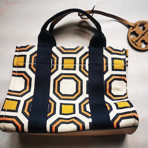 May include: A canvas tote bag with a geometric pattern in white, navy, orange, and yellow. The bag has navy handles and a leather tag with a logo. The bag is approximately 25 cm wide and 20 cm tall.