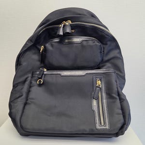 May include: Black nylon backpack with multiple zipped compartments. Features gold-toned hardware, including zips and a small tassel detail. The top pocket is labelled "SUNGLASSES," and the lower pocket reads "IMPORTANT THINGS."