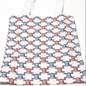 May include: White tote bag featuring a repeating pattern of red and blue interlocking shapes. The bag has white handles and is made of a light-coloured fabric. The design covers the entire front of the bag.