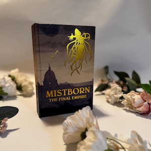 Puede incluir: Un libro de tapa dura titulado "Mistborn: The Final Empire" con una cubierta azul oscuro y letras e ilustraciones doradas. La portada presenta una silueta de una ciudad y una llave. Flores blancas y rosas rodean el libro.