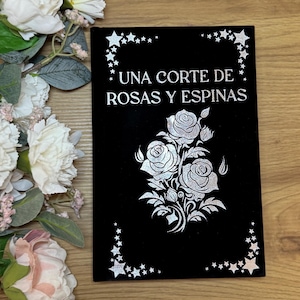 Op de afbeelding: Een zwarte boekomslag met de tekst "UNA CORTE DE ROSAS Y ESPINAS" in het wit. De omslag heeft een zilveren rozenontwerp en steraccenten. Witte bloemen en groen zijn op de achtergrond.