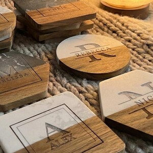 May include: A collection of personalized coasters with a two-tone design. The coasters feature a combination of wood and marble or stone, with engraved names and initials. The shapes include round, square, and hexagonal.