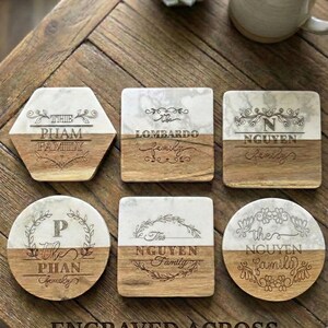 Puede incluir: Una colección de posavasos personalizados con diseño de mármol y madera. Cada posavasos presenta nombres de familia grabados y elementos decorativos. Los posavasos tienen varias formas, incluyendo hexagonal, cuadrada y redonda. También se ve el texto "ENGRAVED ACROSS MARBLE & WOOD".