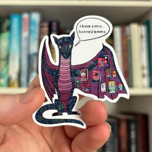 May include: A sticker featuring a cartoon dragon with a purple and blue color scheme. The dragon has a speech bubble that says "I have some... banned books." The dragon's wings are adorned with images of book covers.