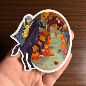 May include: An oval sticker depicting a black horse rearing, with a figure in a blue cloak and yellow ducklings. The background shows a covered bridge and autumn foliage in orange, red, and green hues.