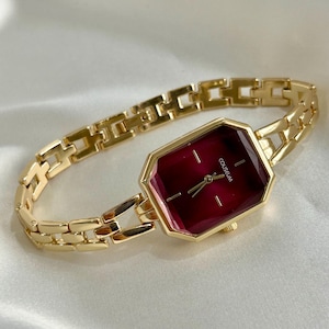 May include: A gold-toned wristwatch with a rectangular, burgundy-colored face and a matching gold link band. The watch face has the word "COLISEUM" printed on it. The watch is displayed on a white, satin-like surface.