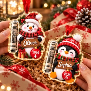 May include: Two clear acrylic Christmas ornaments featuring a snowman and a penguin. The snowman holds a sign reading Oliver, and the penguin holds a sign reading Sophia. Both include a rolled dollar bill, a red hat, and a 2026 ornament.