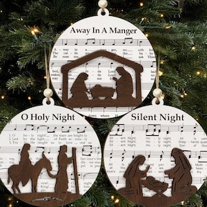 Puede incluir: Tres adornos navideños de madera con temas religiosos. Cada adorno presenta un villancico diferente, con la letra y las notas musicales impresas en la parte delantera. Los adornos tienen un recorte de madera de una escena de la Natividad.