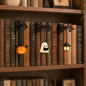 May include: Three black metal wall-mounted brackets with ornate scrollwork, each featuring a hanging light fixture. The lights include an orange jack-o'-lantern with bat wings, a white ghost reading a book, and a classic black lantern with a warm glow.