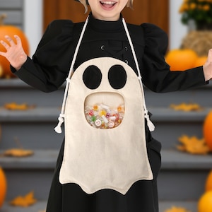 May include: A cream-colored ghost-shaped candy bag with black eye cutouts and a clear window filled with colorful candies. The bag is suspended by a white rope. The image has a Halloween theme.