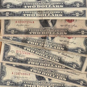 May include: Several United States two-dollar bills are stacked, showing the portrait of Thomas Jefferson. The bills are from the 1963 series and feature the text "Two Dollars" prominently. The bills have serial numbers and the text "United States of America".