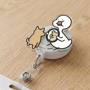 Puede incluir: Un portatarjetas retráctil blanco y transparente con un pato de dibujos animados sosteniendo un espejo con el reflejo de un perro. Un perro marrón está a la izquierda. El portatarjetas tiene una correa de plástico transparente y un tarjetero blanco.