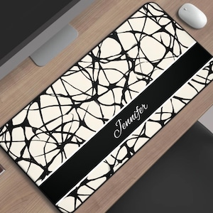 May include: Rectangular desk mat with a cream background and black abstract lines. A black stripe across the mat features the name "Jennifer" in white script. A computer mouse and coffee cup are visible.