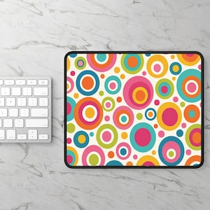 May include: A rectangular mouse pad featuring a repeating pattern of concentric circles in vibrant shades of pink, orange, yellow, teal, and lime green on a white background, with a black stitched border edge and a white computer keyboard.