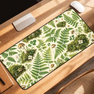May include: Rectangular desk mat featuring a botanical design with green ferns, mushrooms, and white flowers on a cream background. The mat has a black border and is placed on a wooden desk. A computer mouse and a cup of coffee are also visible.