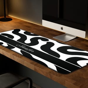 May include: A black and white desk mat with a bold, abstract pattern. A black stripe across the center displays the name "Jennifer" in cursive. The mat is rectangular and rests on a wooden desk, near a computer.