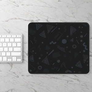 May include: Black rectangular mousepad with a dark gray geometric pattern of triangles, circles, and squiggly lines. A white keyboard is visible on the left side of the image. The mousepad has a black stitched edge.