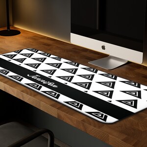 May include: A rectangular desk mat with a black and white geometric triangle pattern. A black stripe runs across the center, with the name "Jennifer" in white script. The mat is on a wooden desk.