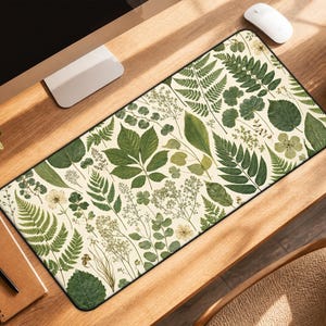 May include: Rectangular desk mat featuring a botanical design with various green leaves and floral elements on a cream background. The mat has a black border and is placed on a wooden desk. A computer, mouse, and coffee cup are also visible.