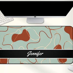 May include: A rectangular desk mat with a teal background featuring rust-colored organic shapes and gold-colored lines. A black band at the bottom displays the name "Jennifer" in white script. A computer, keyboard, and mouse are also visible.