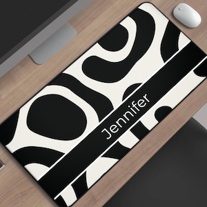 May include: A rectangular desk mat with a black and white abstract pattern. A black stripe across the mat displays the name "Jennifer" in white. A computer mouse and a cup of coffee are visible.