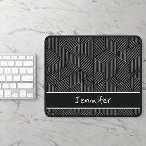 May include: Black and white geometric pattern mousepad with the name "Jennifer" in white text on a black banner. A white keyboard is visible on the left side of the image. The mousepad has a rectangular shape with rounded corners.