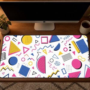 May include: A rectangular desk mat with a white background and a colorful 80s-inspired geometric pattern. The design includes pink, yellow, blue, and black shapes such as triangles, circles, and squares. The mat is placed on a wooden desk.