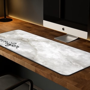 May include: A rectangular desk mat with a white marble design and the name "Jennifer" in black script. The mat is placed on a wooden desk, with a computer monitor in the background.