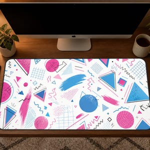 May include: A rectangular mouse pad with a white background and a colorful 80s-inspired geometric pattern. The design includes pink, blue, and black shapes such as triangles, circles, and brushstrokes. The mouse pad is on a wooden desk.