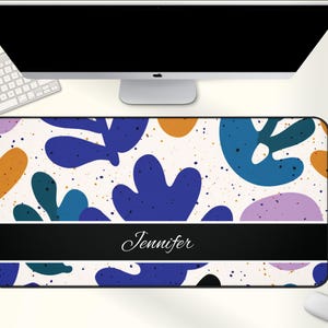 May include: A rectangular desk mat with a white background and a colorful abstract design. The design includes blue, orange, and purple shapes with gold speckles. A black band at the bottom displays the name "Jennifer" in white script.