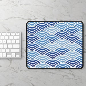 May include: A rectangular mouse pad featuring a repeating pattern of concentric semicircles in various shades of blue and white. The design consists of layered, wave-like arcs arranged in a grid, finished with a solid black stitched border edge.