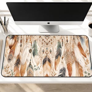 May include: A rectangular desk mat featuring a watercolor design of feathers in shades of brown, beige, and green. The mat has a black border and a macrame-style design at the top. The desk mat is placed on a white desk.