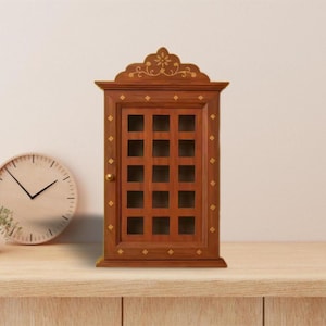 Puede incluir: Un mueble para llaves de madera con un diseño de puerta enrejada. El mueble es de color marrón cálido, con detalles dorados y una parte superior decorativa. También se ven un reloj redondo y una pequeña planta en maceta.