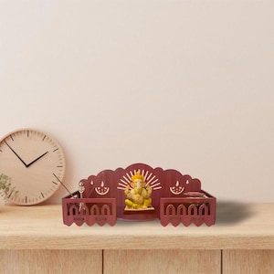 Könnte beinhalten: Ein weinroter Wandorganizer aus Holz mit einem mittigen Ausschnitt für eine goldene Statue von Ganesha. Der Organizer hat zwei Seitenfächer und dekorative Ausschnitte. Eine Holzuhr und eine kleine Pflanze befinden sich links.
