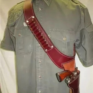 Può includere: Una fondina a tracolla in pelle bordeaux con una cintura di munizioni abbinata, indossata sopra una camicia verde a maniche corte. La fondina contiene un revolver argento e marrone. La cintura di munizioni ha diversi bossoli.